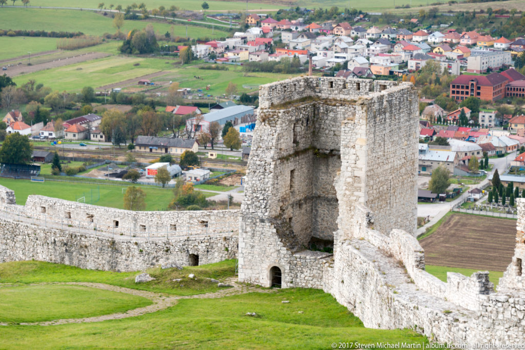 Square ruined stone tower with a small arched opening projecting from a long castle wall above green grass.