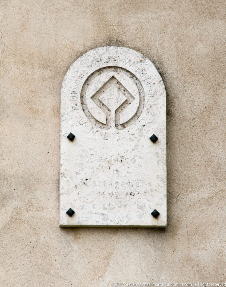 Arched pale plaque with a carved geometric emblem and faint lettering mounted on a beige wall.