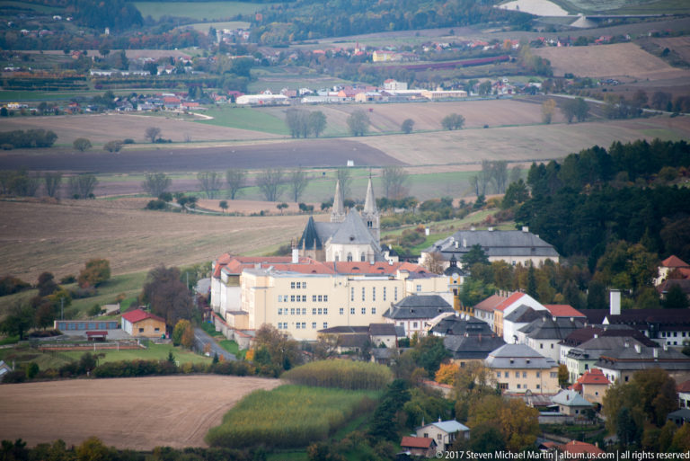 Church with pointed spire and nearby pale building in a village surrounded by patchwork agricultural fields.