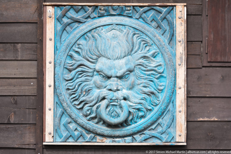 Blue-green relief panel with a bearded face and geometric border mounted on dark wooden boards.