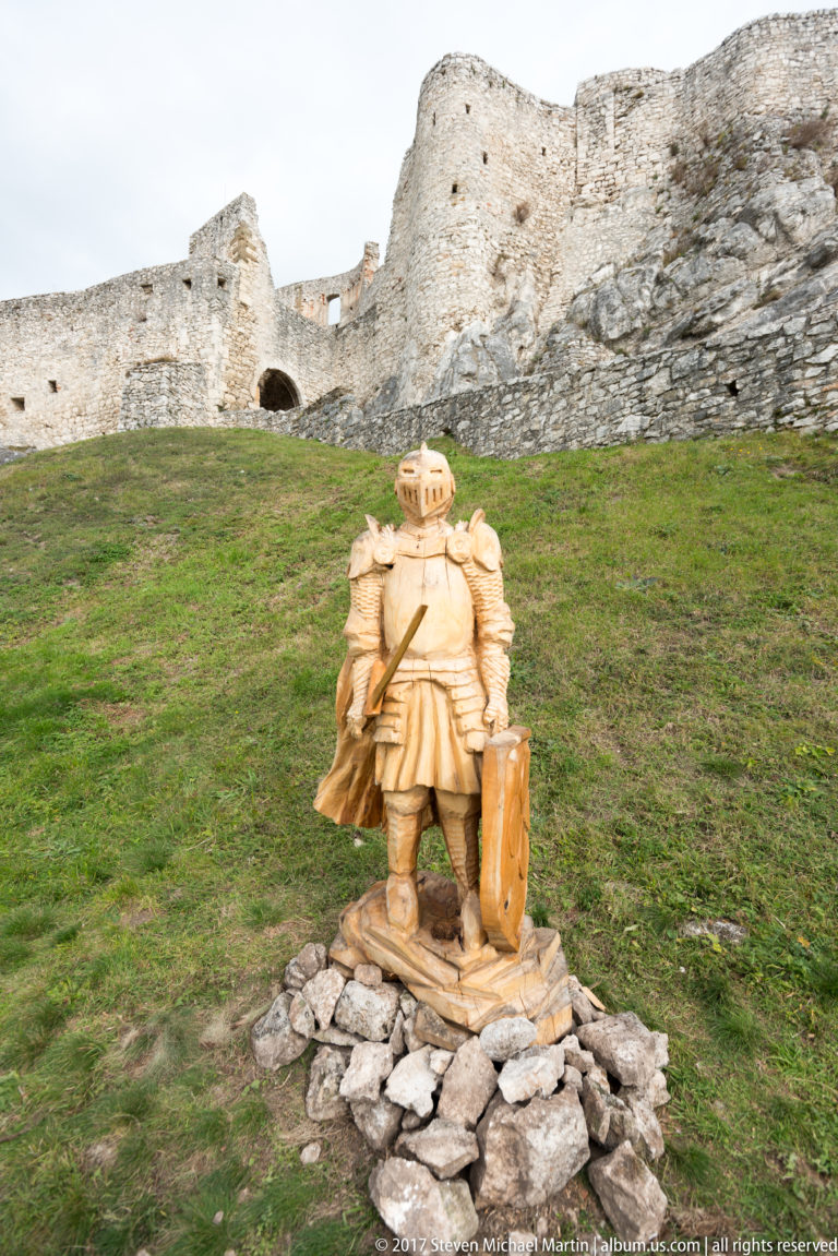 Carved wooden knight with shield standing on stones before ruined castle walls and green grass.