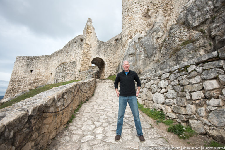 Man standing on a stone path leading toward a ruined arched gateway between castle walls.