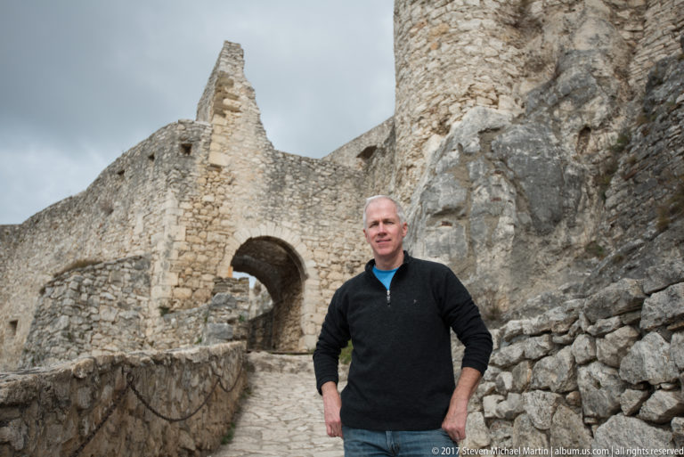 Man standing beside a stone path and arched castle gateway amid rough ruined masonry.