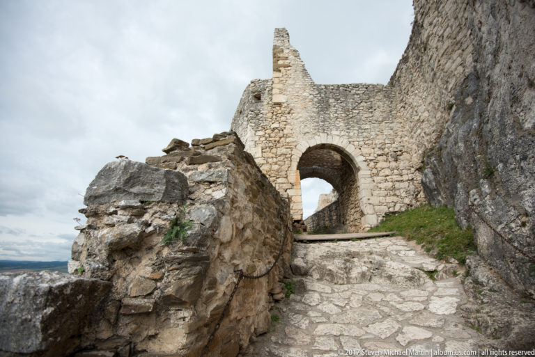 Stone path climbing through a round-arched opening between ruined castle walls and exposed rock.