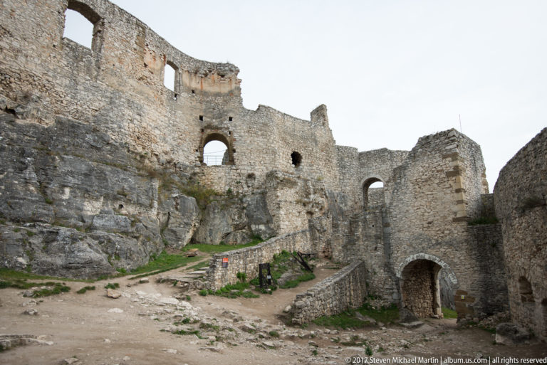 Ruined castle courtyard with arched openings, layered stone walls, paths, and exposed bedrock.