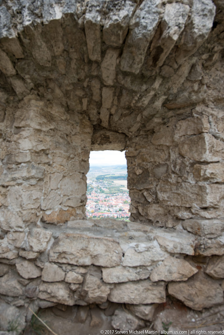Small rectangular opening in a thick stone wall framing fields and settlement far below.