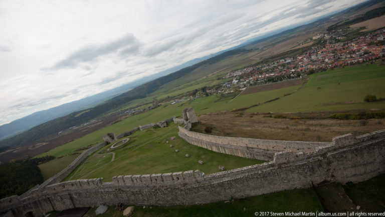 High angled view over Spiš Castle's lower wall and grassy enclosure toward fields and a red-roofed settlement.