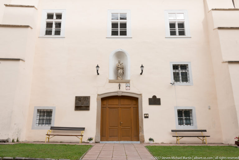 Pale historic façade with central wooden door, niche statue, wall plaques, benches, and rectangular windows.