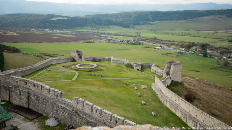 Elevated view of a grassy castle ward enclosed by stone walls and towers, with fields beyond.