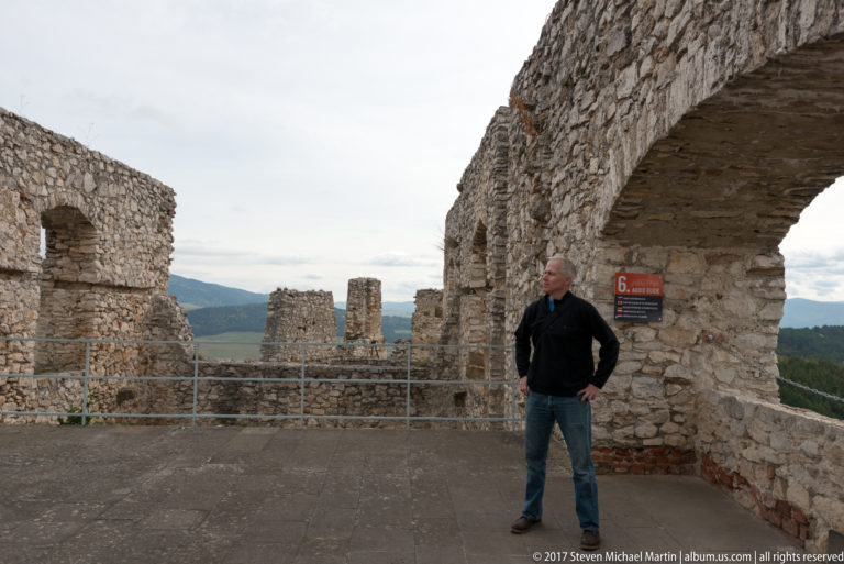 Man standing beneath a broad stone arch amid castle ruins, railings, and a red interpretive sign.