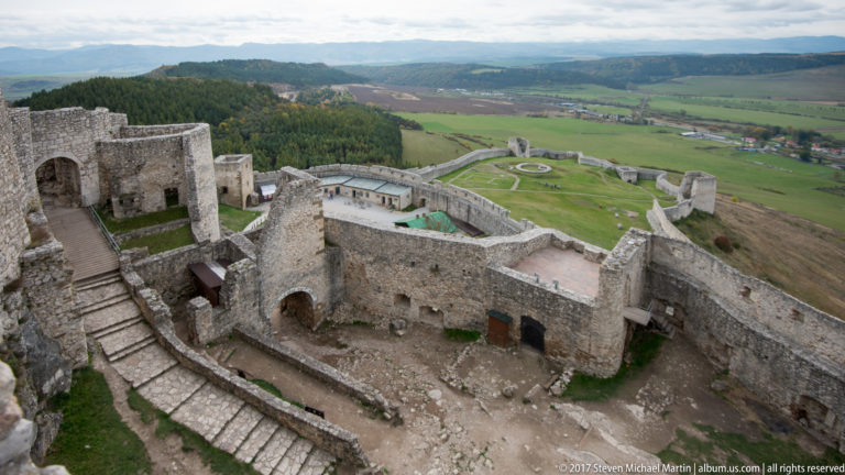 Elevated view over stone stairs, ruined rooms, walls, and the lower castle enclosure extending into the landscape.