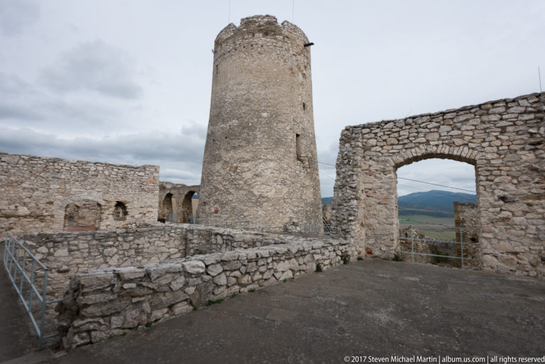 Cylindrical stone tower beside ruined walls and a large rectangular opening framing distant hills.