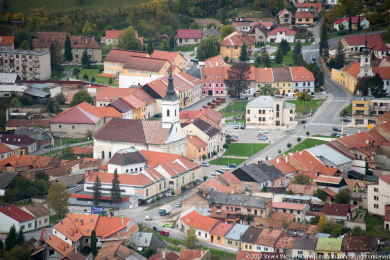 White church with dark spire surrounded by red-roofed buildings and streets in the town below the castle.