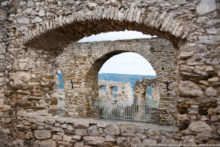 Two aligned stone arches in ruined walls framing distant hills, with a metal railing across the inner opening.