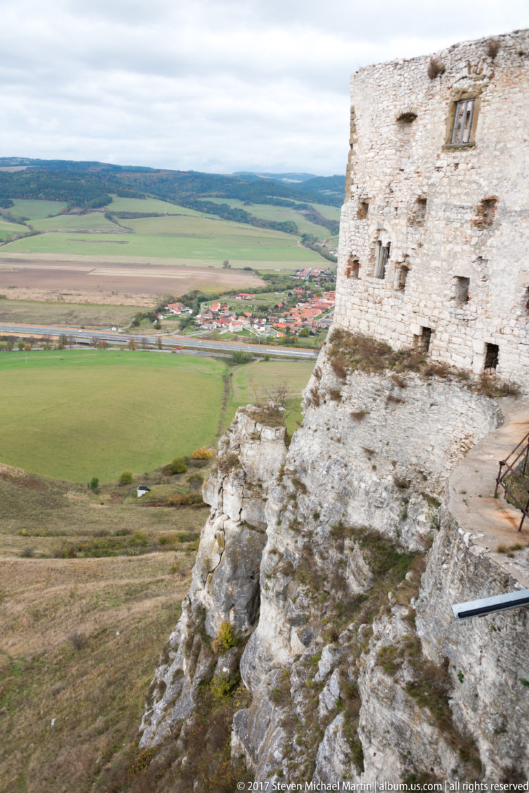 Ruined castle wall perched on exposed rock above broad fields and a distant settlement.
