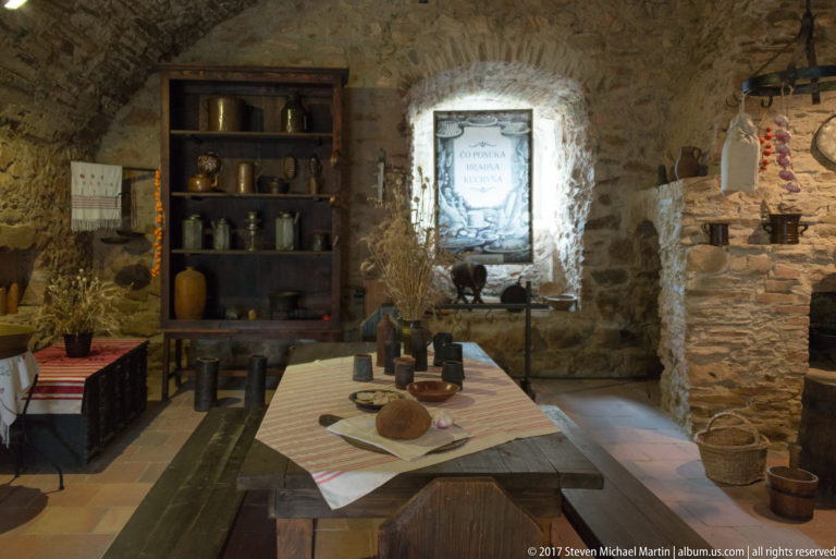 Reconstructed stone room with wooden table, pottery shelves, baskets, food display, and small window.