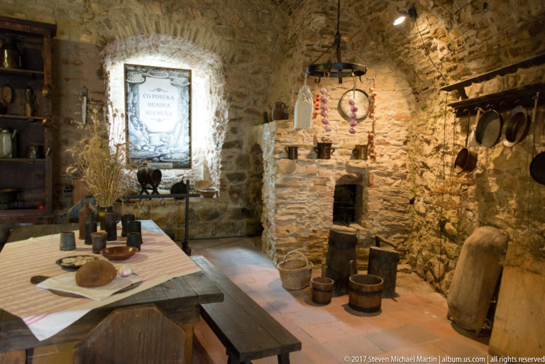 Reconstructed castle kitchen with masonry hearth, hanging pans, baskets, wooden vessels, and a set table.