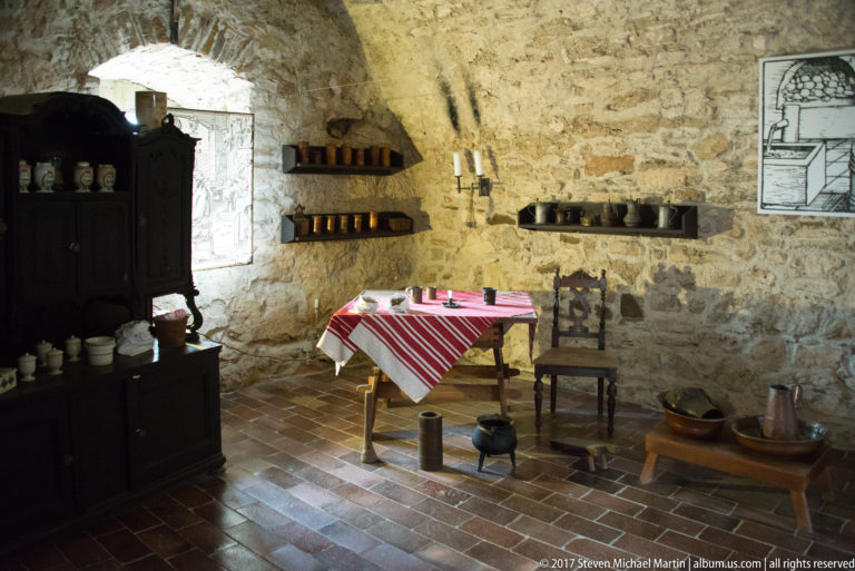 Stone room arranged with striped table, ceramics, wall shelves, chair, cupboard, and household vessels.