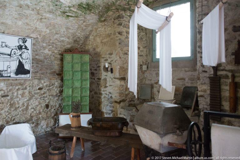 Stone room with large metal basin, green tiled structure, hanging white linen, bench, and utility equipment.