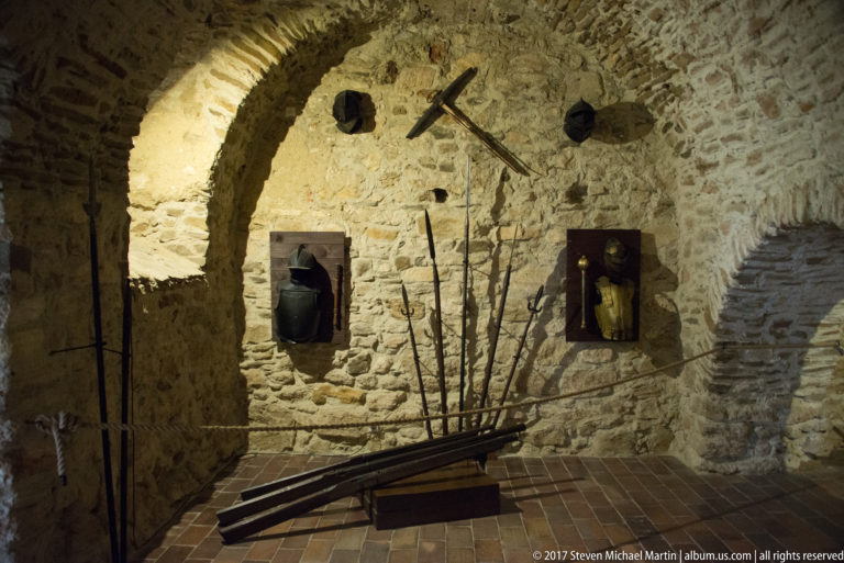 Long polearms and staffs displayed against a rough stone wall in a vaulted castle chamber.