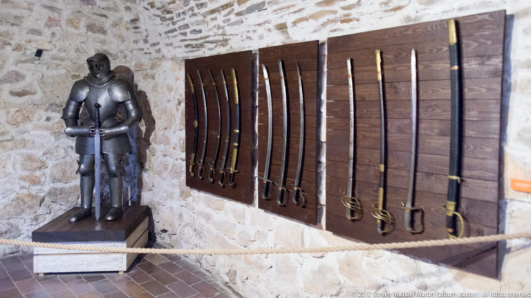 Full suit of dark plate armor beside multiple swords mounted vertically on wooden panels.