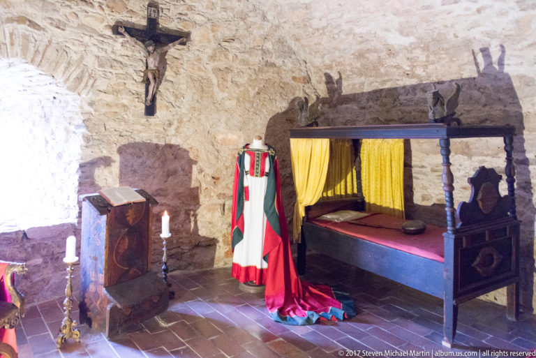 Canopied wooden bed with yellow curtains beside a red-and-green historic costume, chest, and candlesticks.