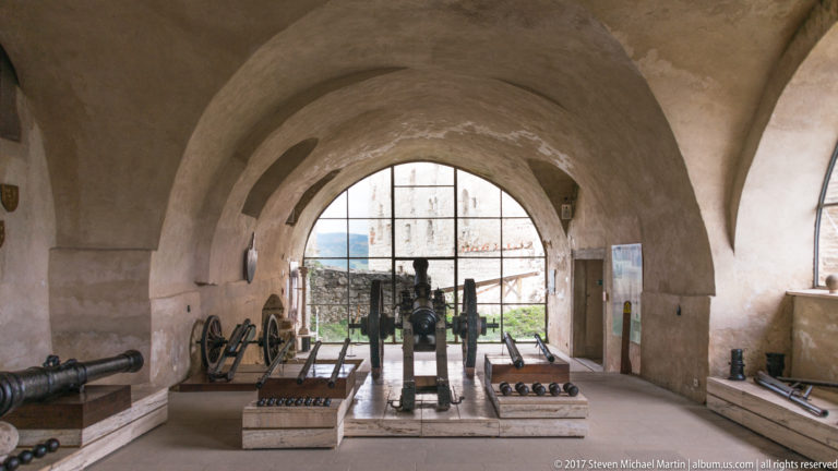 Historic cannons arranged in a vaulted castle gallery with a large arched opening at the far end.