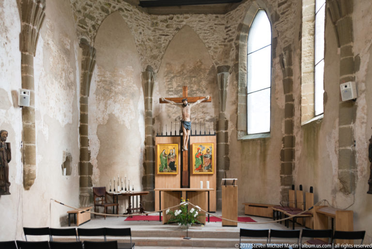 Stone chapel with large crucifix, devotional panels, altar, pointed windows, chairs, and lectern.