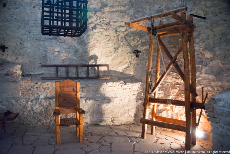 Wooden chair beside a tall timber restraint-like apparatus, ladder, and hanging cage in a stone chamber.
