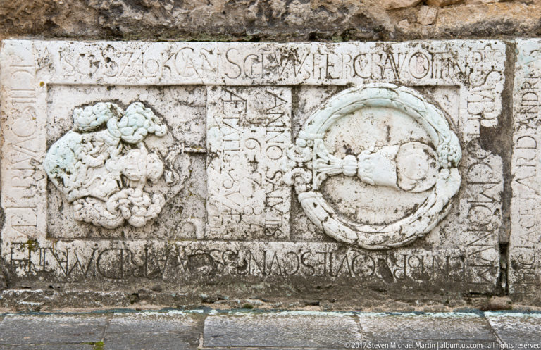 Weathered rectangular stone relief with two heraldic shields, a small central shield, and worn carved lettering.