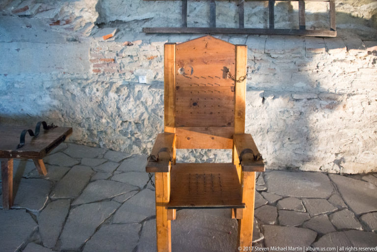 Heavy high-backed wooden chair with broad armrests standing against a rough stone wall.