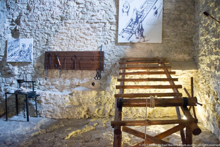 Wooden rack-like restraint apparatus with chains and iron tools displayed in a rough stone chamber.