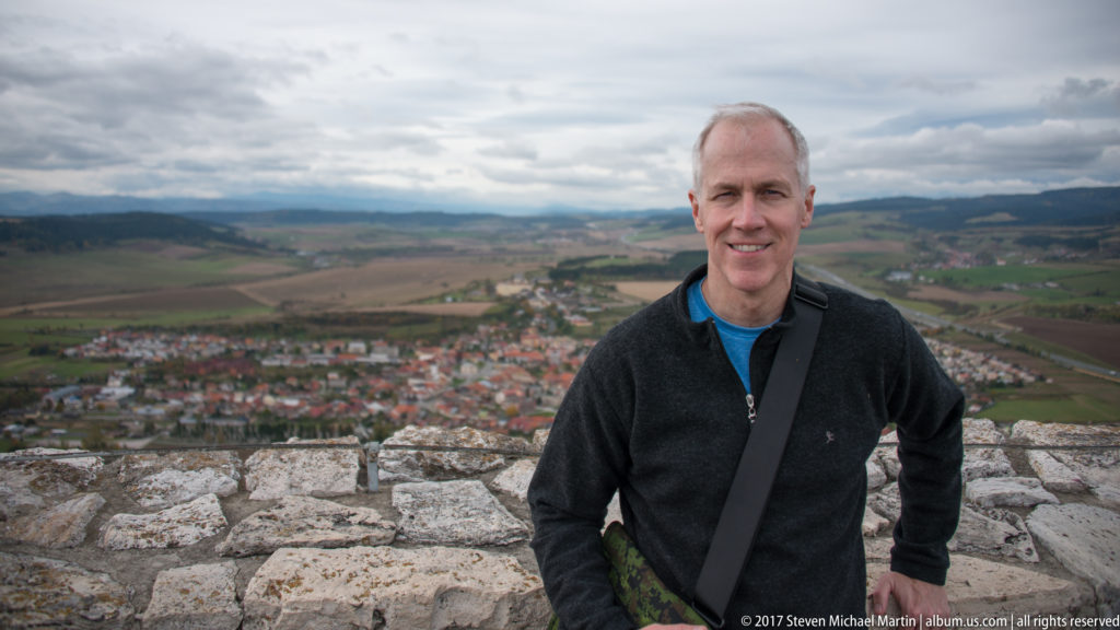 Man standing on stone castle walls with red-roofed town, fields, and low hills in the background.