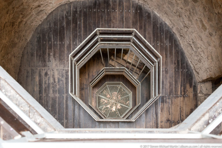 Downward view into an octagonal timber-lined shaft with repeated railings and a star-patterned panel at the bottom.