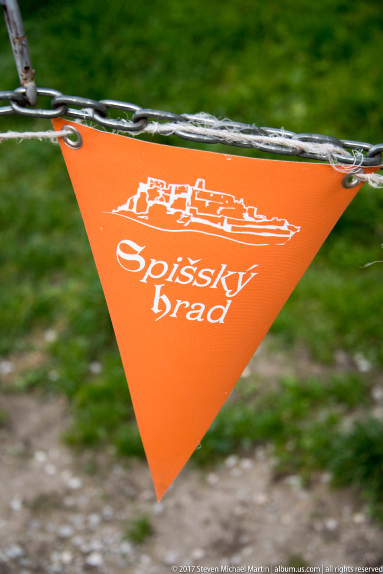 Orange triangular pennant on a chain printed with a white castle silhouette and the words “Spišský hrad.”