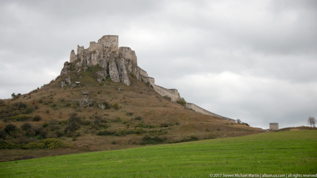 Spiš Castle ruins on a steep rocky hill above a green field beneath a cloudy gray sky.