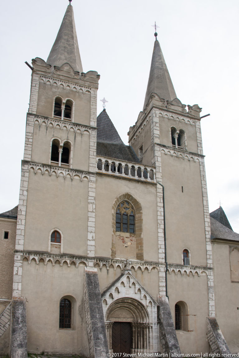 Twin-towered medieval church façade with recessed portal, traceried window, arcaded bands, and steep roofs.