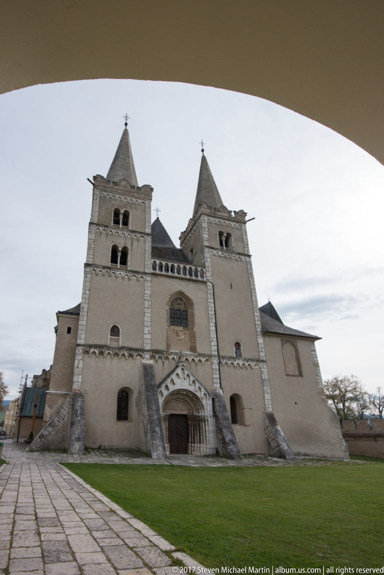 Wide view through an arch toward a twin-towered medieval church across a paved and grassy courtyard.