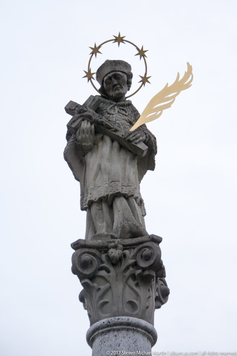 Stone saint statue with halo of stars and gilded branch-like attribute atop an ornate column.