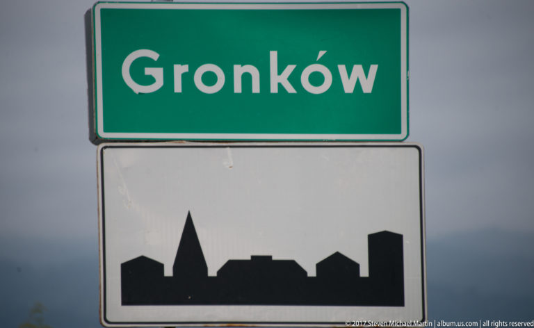 Green Gronków road sign above a black built-up-area silhouette beneath a dark cloudy sky.