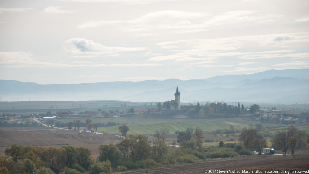 Distant village church tower among fields and trees, with hazy blue-gray mountain ridges layered across the horizon.