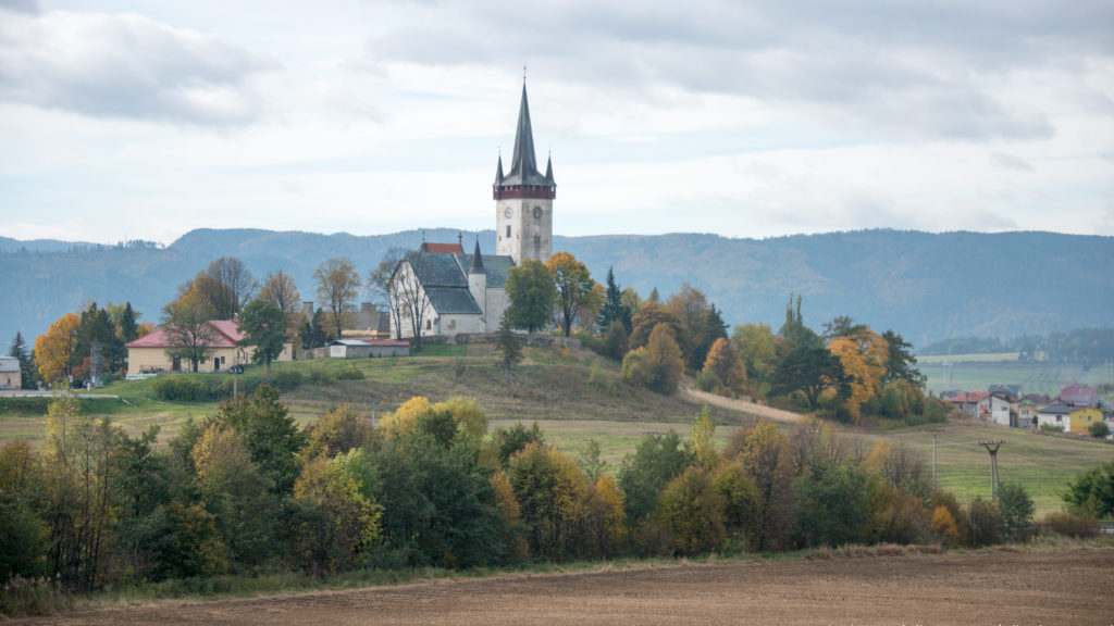 Twin-spired hilltop church surrounded by autumn trees and village houses, with fields and a wooded ridge beyond.