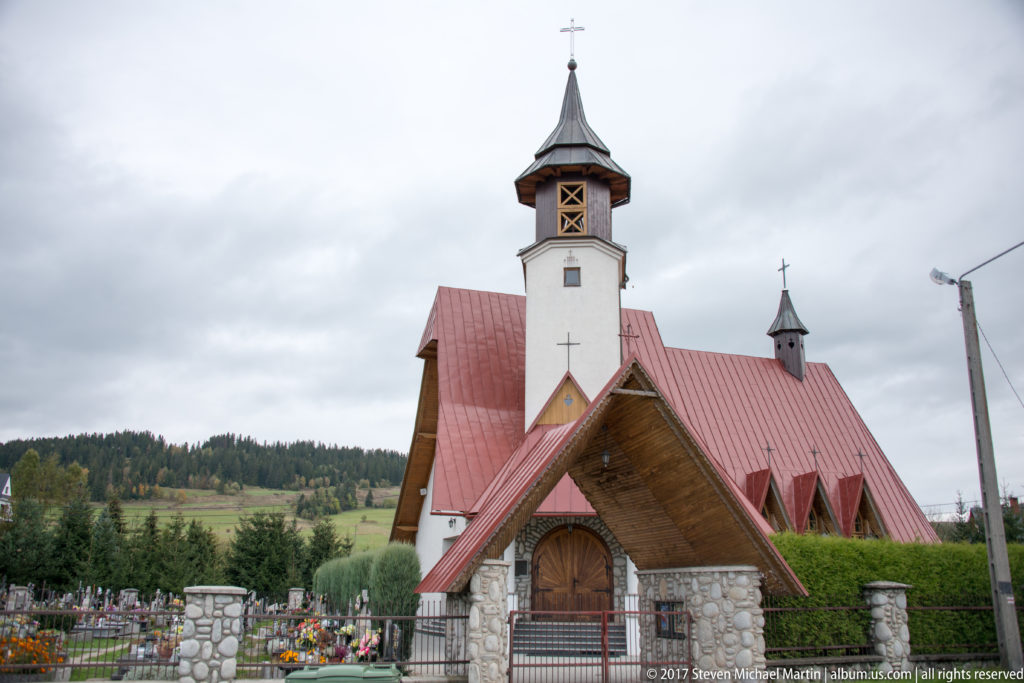 Stone village church with red roof, timber entrance canopy, bell tower, and flower-filled cemetery in the Tatra foothills.