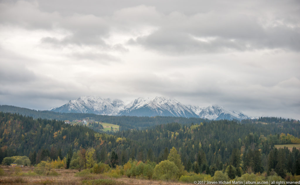 Forested Tatra foothills with autumn foliage and pale mountain peaks beneath a heavy gray cloud deck.