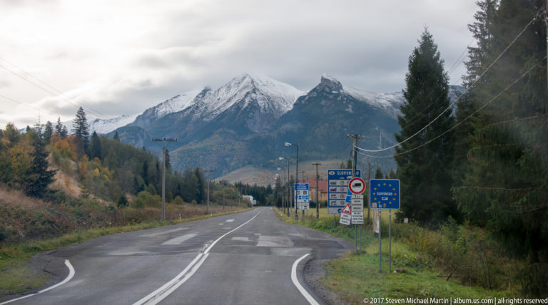 Two-lane mountain road leading toward rocky Tatra peaks and roadside signs near the Slovak border.