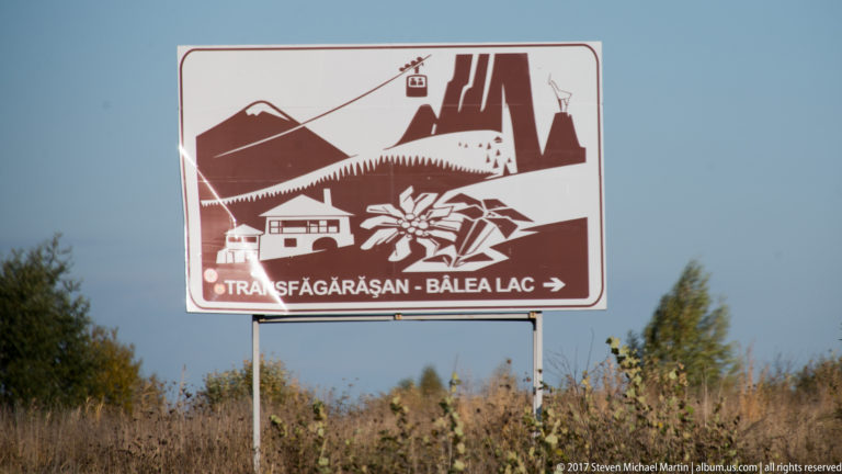 Brown-and-white Transfăgărășan–Bâlea Lac directional sign above roadside vegetation under a clear blue sky.