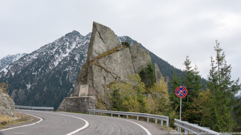 Tall angular rock pinnacle beside a curving mountain road, guardrail, and snow-dusted ridge.