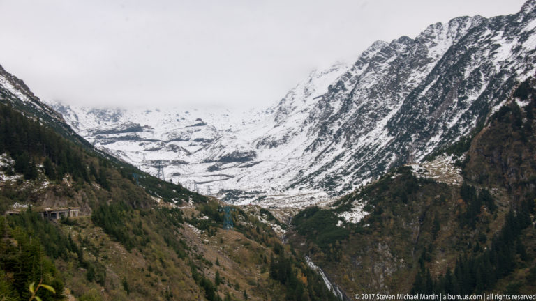 Steep snow-covered alpine valley with dark rock ribs, conifers, and road structures along the lower slope.