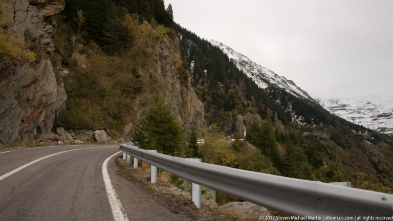Mountain road curving between rock wall and guardrail toward a snow-covered high valley.