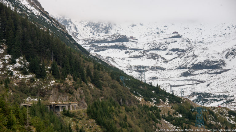 Snowy mountain basin with Transfăgărășan switchbacks, electrical pylons, and a protective road gallery.